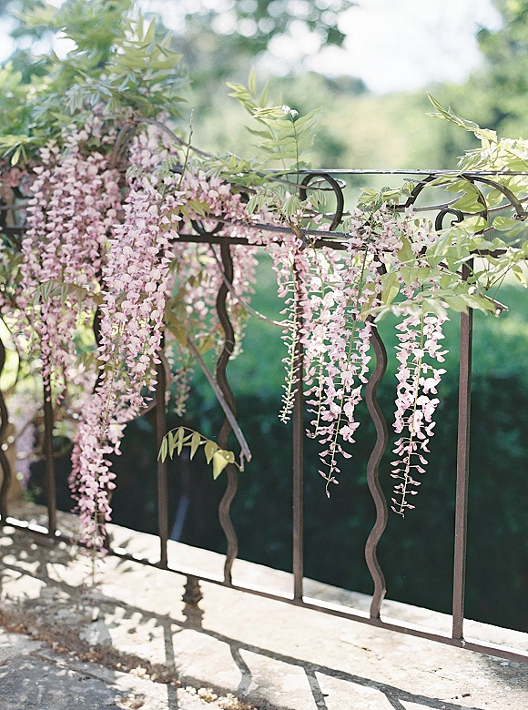 Wisteria decor draped along a wrought iron railing, with cascading wisteria blooms and greenery vines over a stone ledge outdoors