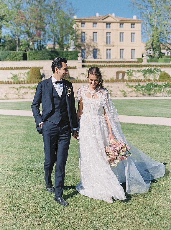 Couple portrait of bride and groom walking hand in hand on an estate lawn, her long veil blowing as he looks at her