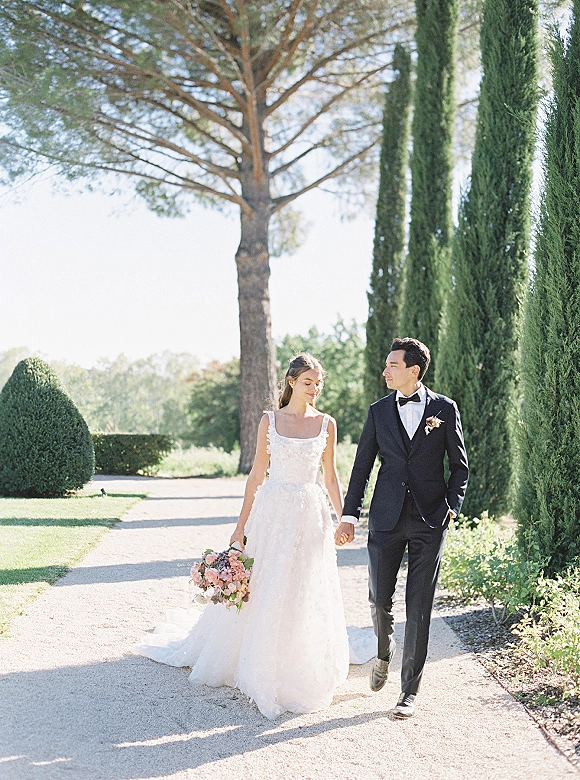 Couple portrait of bride and groom walking hand in hand, bride holding bouquet with veil flowing on a gravel path by cypress trees