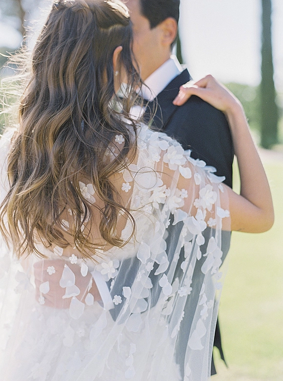 Wedding couple portrait of bride and groom embrace, kissing in sunlight, her floral lace veil and backless gown framed by greenery