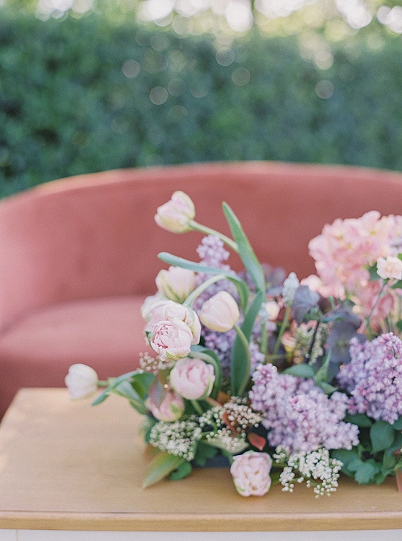Wedding floral arrangement garden wedding centerpiece of pink tulips, lilac blooms, and eucalyptus on a table beside a terracotta velvet sofa outdoors