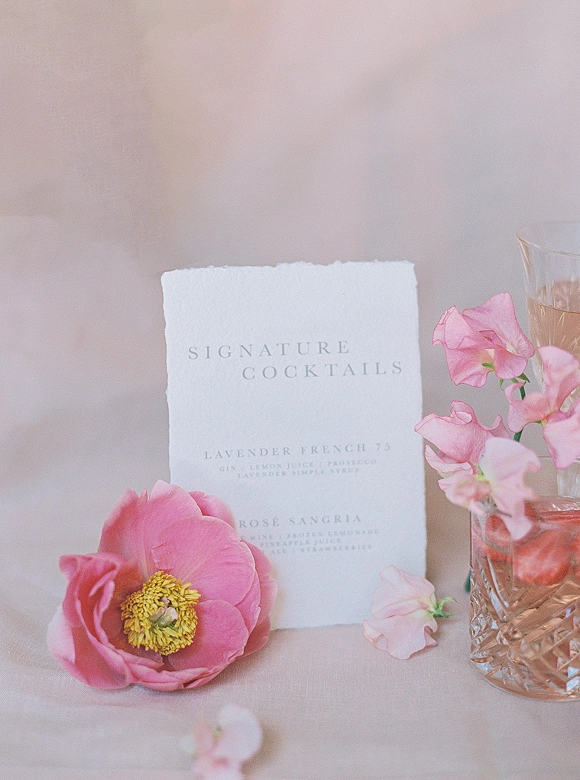 Signature cocktail sign and wedding signature cocktail menu on deckle-edge paper beside a rosé drink with ice and pink peony on linen tablecloth