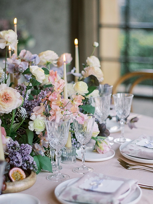 Reception tablescape with wedding table centerpiece of pastel blooms, taper candles, crystal goblets, and place cards in window-lit dining room