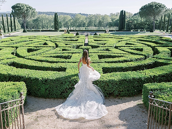 Bridal portrait of a bride from behind holding her lace wedding dress train by an iron railing in a formal hedge maze garden path