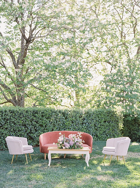Outdoor lounge setup with a velvet sofa and upholstered chairs around a coffee table topped with a floral centerpiece on a garden lawn