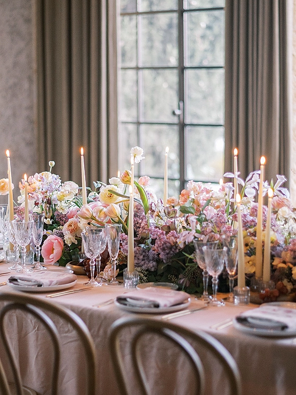 Reception tablescape with wedding head table flowers, pastel centerpiece, taper candles and glassware on neutral linens by a sunlit window