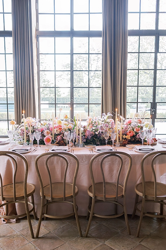 Reception tablescape with pastel wedding centerpiece, taper candles, crystal stemware and white plates on a blush cloth near grid windows.