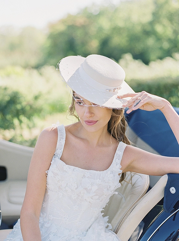Bridal portrait of a bride wearing hat with a birdcage veil, seated in a vintage convertible, looking away in soft greenery backdrop