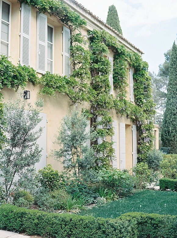 Wedding venue exterior with flowering climbing vines on a stucco villa, white shutter windows and lantern, bordered by manicured hedges and lawn