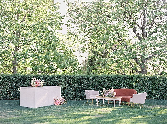 Wedding lounge area with an outdoor wedding lounge sofa, upholstered chairs and coffee table beside a bar on a garden lawn with hedge and trees
