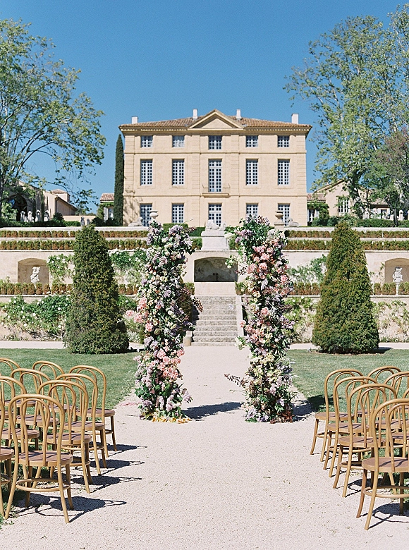 Outdoor ceremony setup with floral ceremony pillars framing a gravel aisle lined with wooden chairs in a terraced manor garden under blue sky