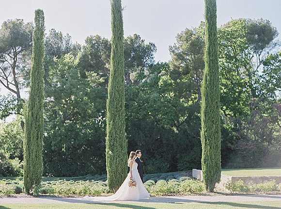 Couple portrait of bride and groom walking side profile, bride with veil and bouquet, groom in tuxedo, by cypress trees and stone wall