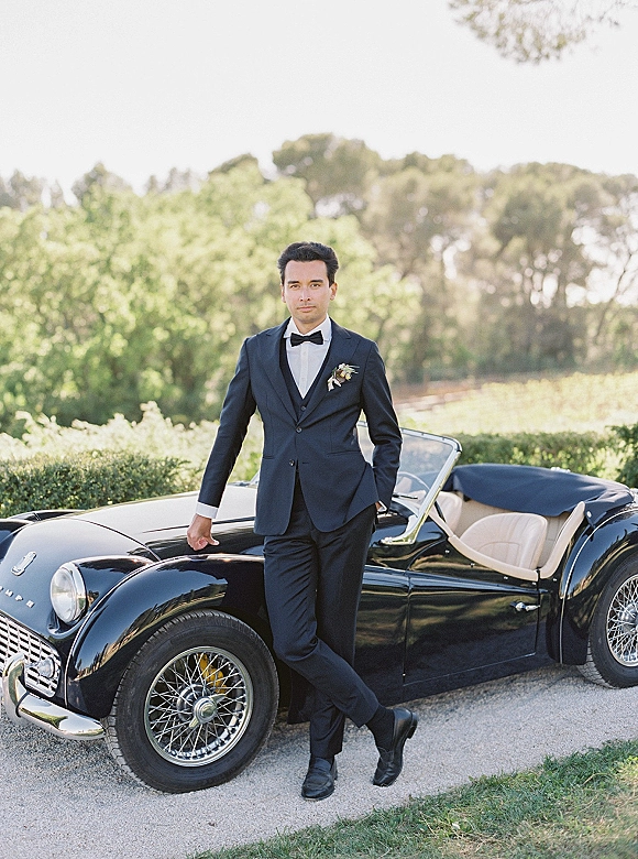 Groom portrait of a groom in tuxedo leaning on a vintage convertible car, wearing a bow tie and boutonniere on a gravel driveway