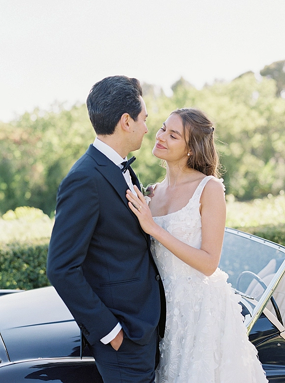 Couple portrait of bride leaning on groom in a black tuxedo beside a vintage car, her lace dress and pearl earrings in soft garden light