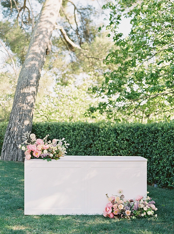 Wedding welcome table with pastel floral arrangements and greenery on a white table, set on a garden lawn with hedges and a large tree in daylight