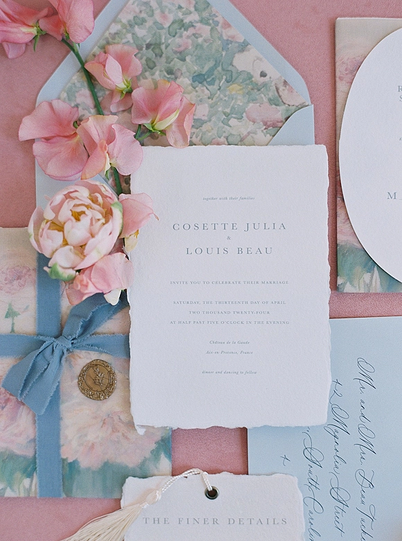 Invitation suite with a deckled edge wedding invitation on pink fabric, featuring floral liner envelope, blue ribbon, wax seal, and tassel place card
