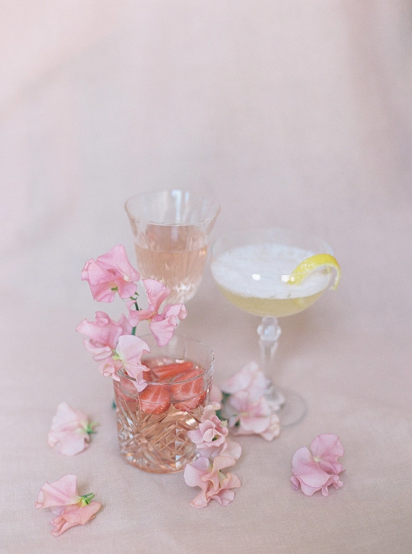 Wedding cocktails trio in coupe, rocks, and wine glasses with pink drink, champagne, strawberry and lemon garnishes, sweet pea accent on blush backdrop