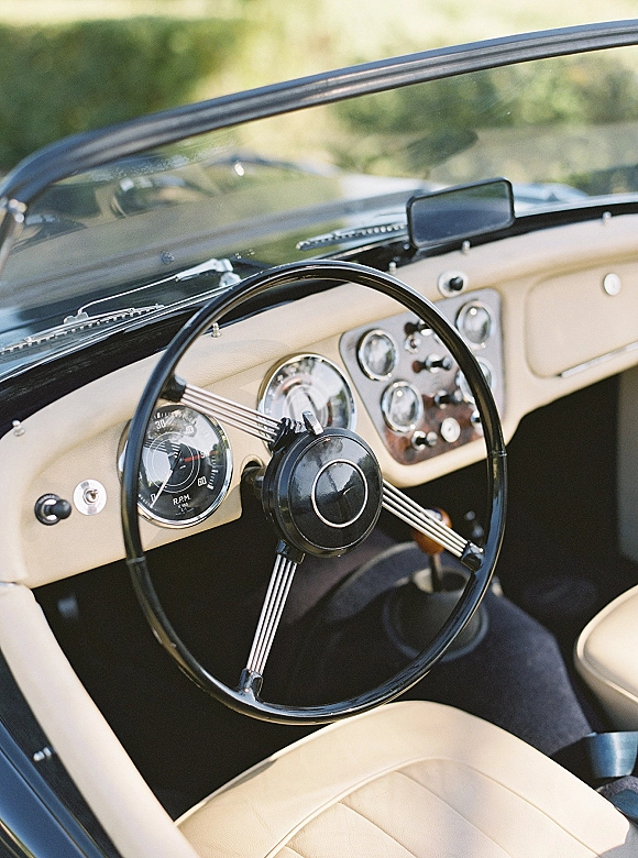 Vintage wedding car interior with classic car wedding details, cream leather seats, steering wheel and gauges glowing in sunlight and greenery