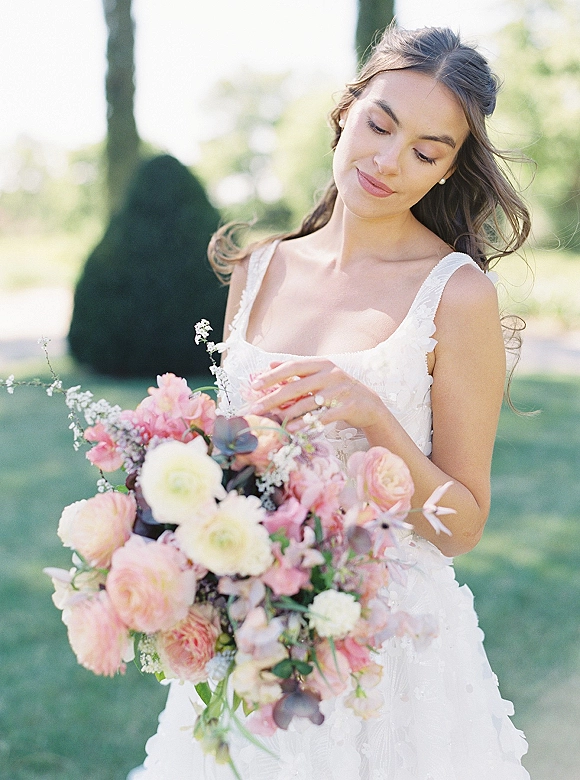 Bridal portrait of a bride holding bouquet, looking down in a sleeveless wedding dress with pearl earrings on a sunlit garden lawn