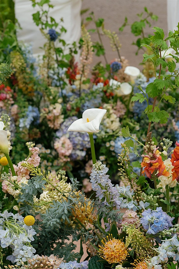 Wedding floral arrangement with calla lilies and hydrangeas, lush greenery and wildflowers set against a neutral wall and draped fabric backdrop