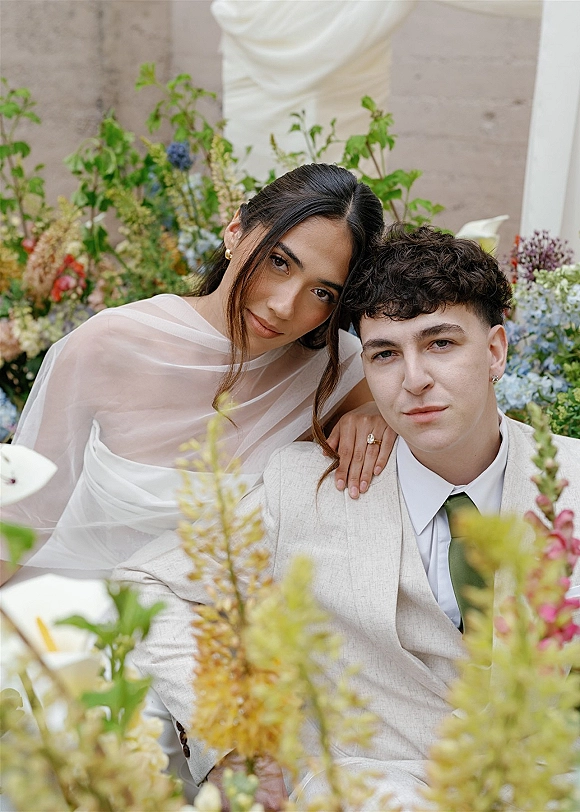 Couple portrait of bride and groom close up, bride leaning on groom in beige suit and veil, framed by soft garden florals.