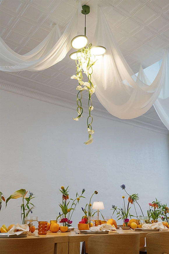 Reception tablescape with citrus wedding tablescape details—wildflower bud vases, amber glassware, and hanging florals beneath draped fabric and pendant light