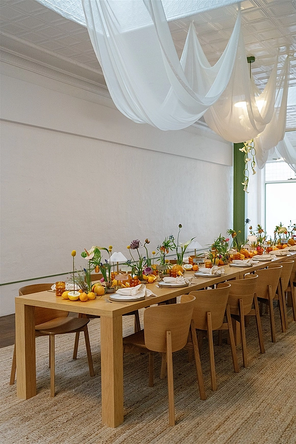 Reception tablescape on a long banquet table with bud vase florals, citrus centerpieces and amber glassware beneath draped ceiling fabric