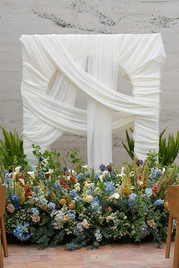 Wedding ceremony backdrop with draped fabric wedding arch, white fabric over greenery and grounded florals by wood chairs on terracotta tile floor