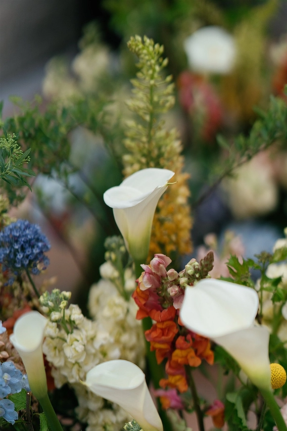 Wedding florals, calla lily arrangement with white blooms, orange and blue flowers, and greenery against blurred garden foliage