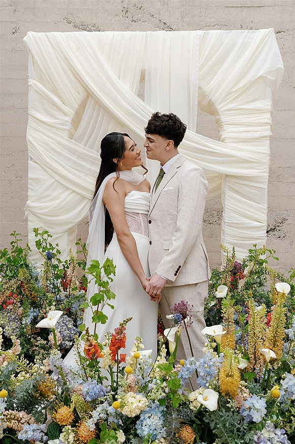 Couple portrait of bride and groom holding hands, smiling under wedding arch draping with wildflower meadow florals against a concrete wall