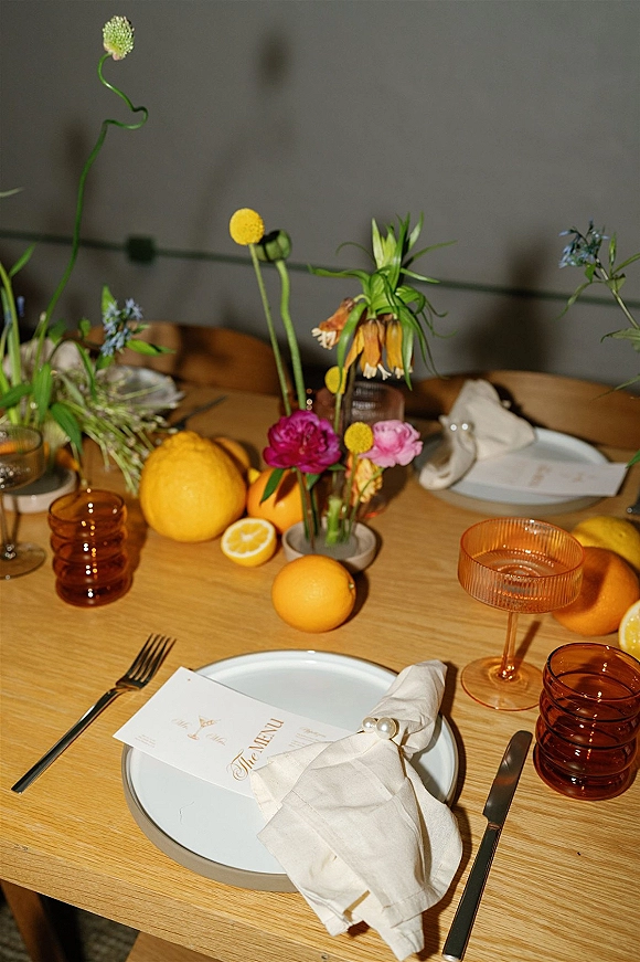 Reception tablescape with citrus wedding tablescape details, bud vase flowers, orange glass coupes, amber tumblers, and pearl napkin rings on a wood table