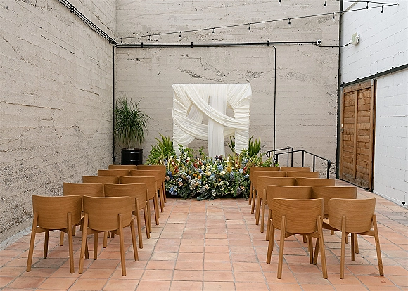 Ceremony setup with a white draped altar and grounded florals, wooden chairs in rows, string lights against concrete walls and terracotta tile floor