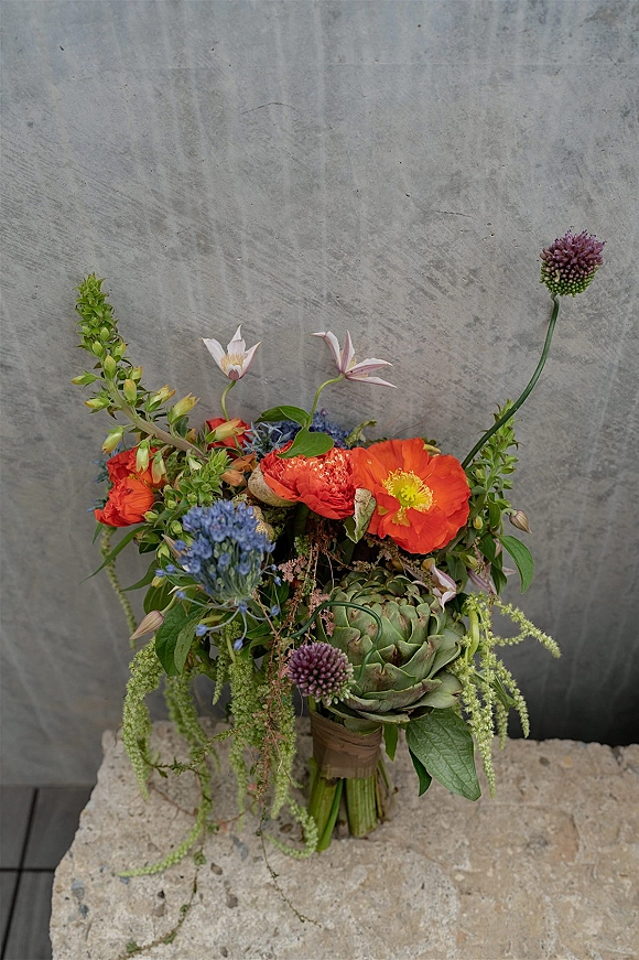 Bridal bouquet with wildflower bridal bouquet styling, featuring orange poppies, succulents, blue blooms and ribbon wrap against a concrete wall
