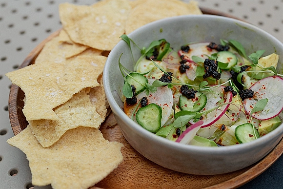 Chip and dip with tortilla chips appetizer in a ceramic bowl on a wooden board, topped with microgreens and chili flakes on a metal table