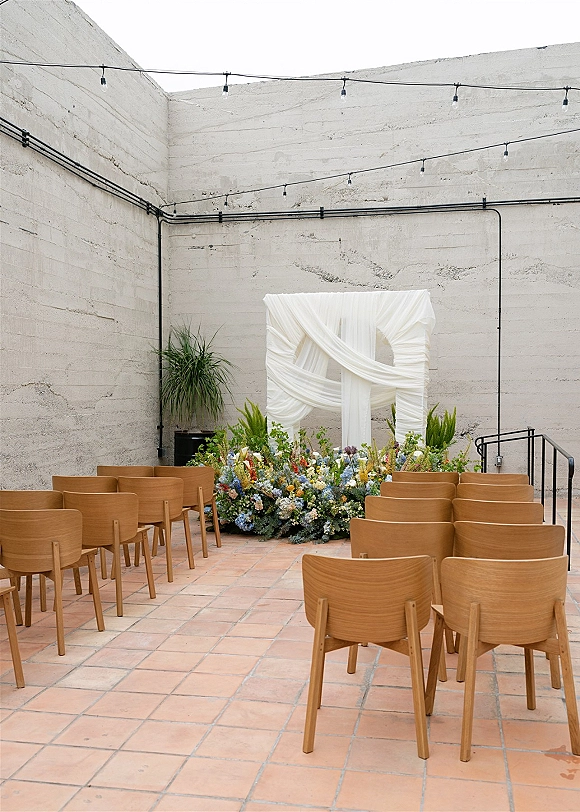 Ceremony setup with a wedding ceremony backdrop, draped fabric and blue-yellow florals, wooden chairs and string lights in an industrial courtyard