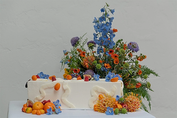 Wedding cake with fresh flowers and citrus fruit, white textured square tier topped with figs and raspberries on a table by a stucco wall