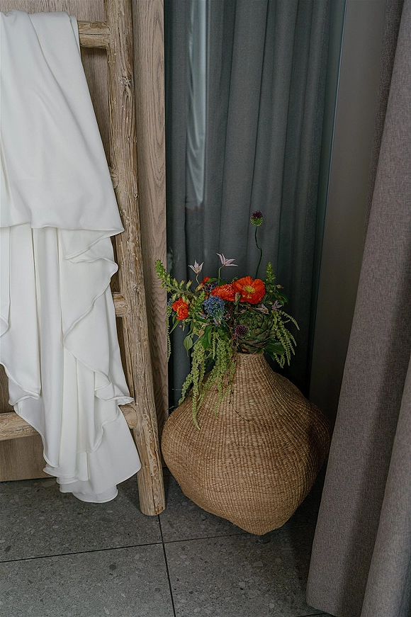Wedding dress detail with ruffle fabric displayed on a rustic wooden ladder beside a woven basket vase of flowers against gray curtains