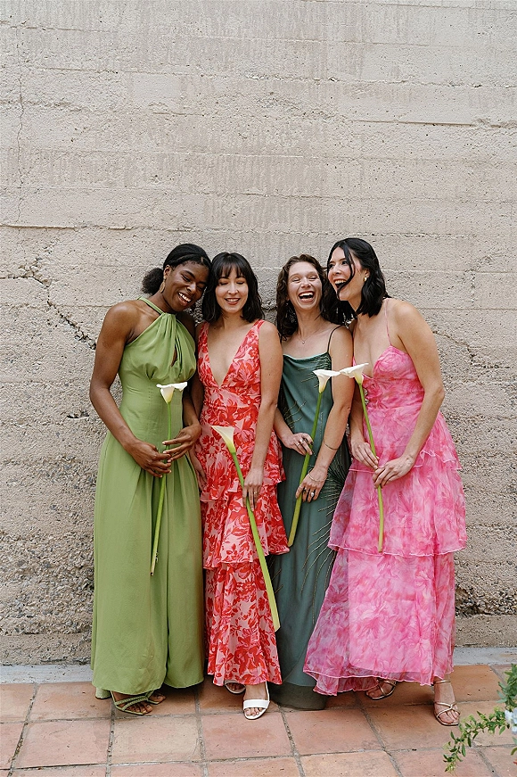 Bridesmaid group photo of women laughing in colorful mismatched dresses, holding single-stem calla lilies by a textured concrete wall