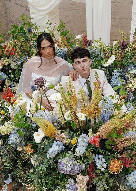 Couple portrait of bride and groom sitting amid calla lilies and blue hydrangeas, bride’s sheer cape against a draped fabric stone wall backdrop