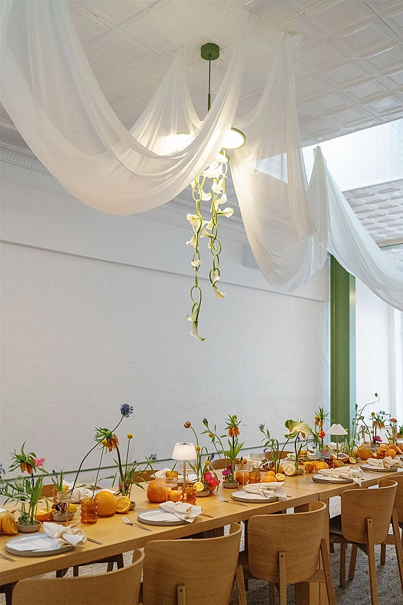 Reception tablescape on a long wooden table with amber glassware, white plates, pumpkins and citrus, under hanging flowers and white drapery