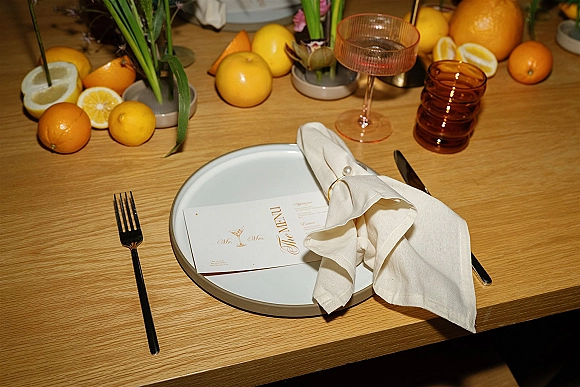 Wedding place setting with wedding menu on plate, linen napkin in a pearl ring, black flatware, amber tumbler, coupe glass on wood table