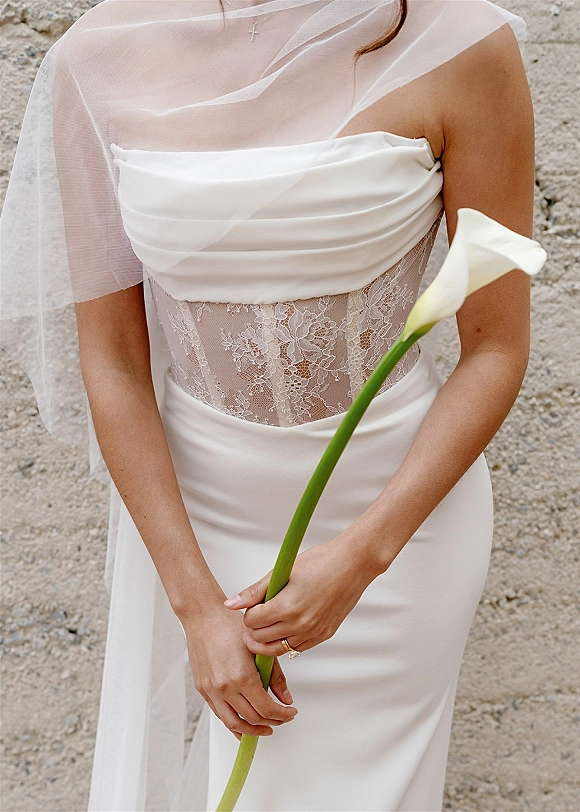 Wedding dress with strapless wedding dress lace bodice, tulle veil and calla lily in hand, engagement ring visible against a stone wall