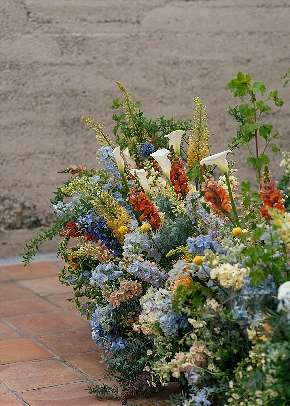 Wedding floral arrangement of calla lilies, delphinium, snapdragons and billy balls with lush greenery on terracotta tiles by a stone wall