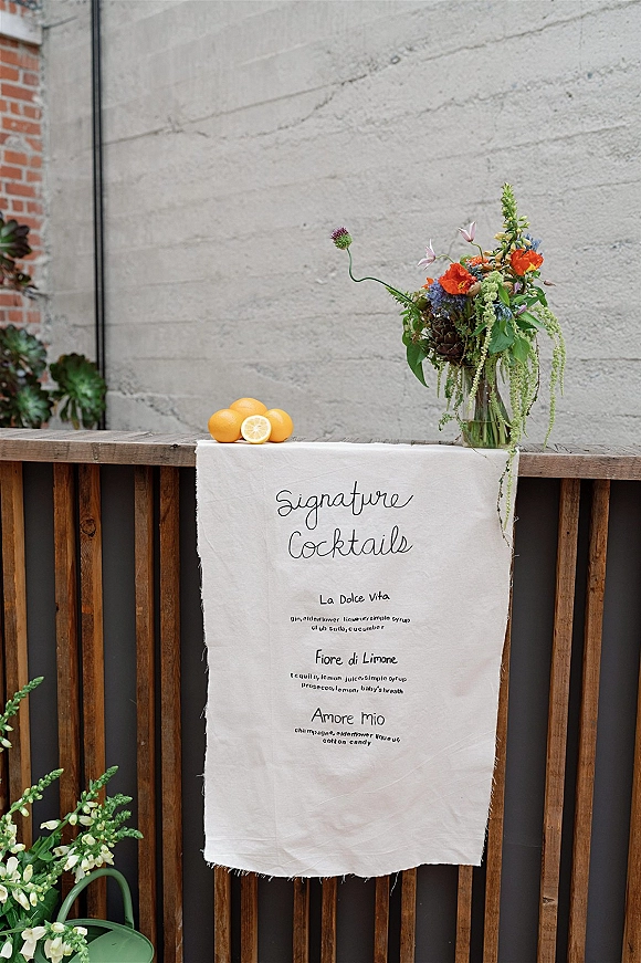 Wedding bar signage on linen fabric with calligraphy cocktail menu, oranges, and floral vase on a wooden bar against concrete and brick walls