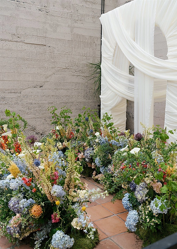 Ceremony aisle decor with meadow aisle flowers, hydrangeas and calla lilies amid greenery and moss, set by a draped fabric backdrop on terracotta tile floor