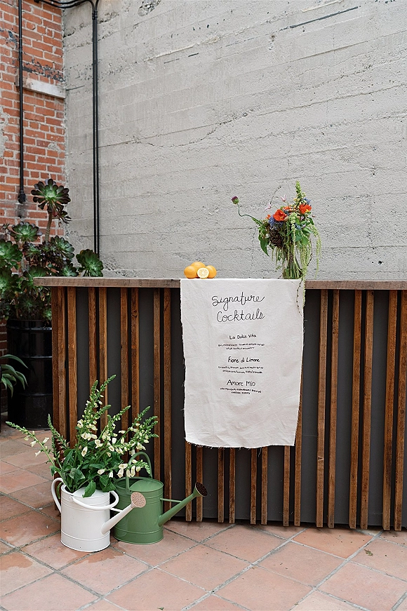 Signature cocktail sign on a linen menu banner with calligraphy, citrus and floral accents on a wood bar against brick and concrete walls