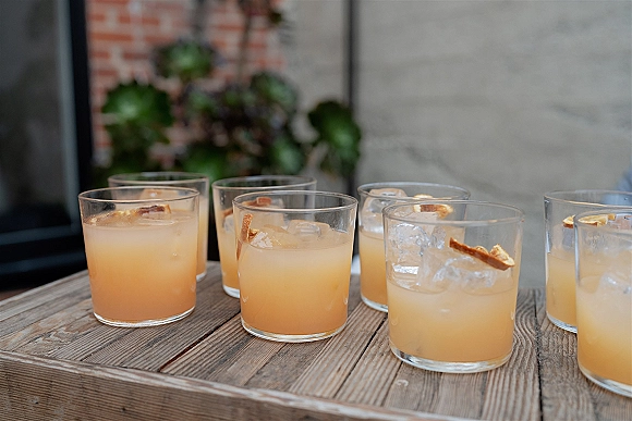Signature wedding cocktails in rocks glasses with ice cubes and citrus garnish on a wood table against a brick wall with greenery