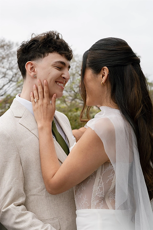 Couple portrait with bride showing engagement ring as she touches groom’s face, lace dress and veil framed by trees under overcast sky