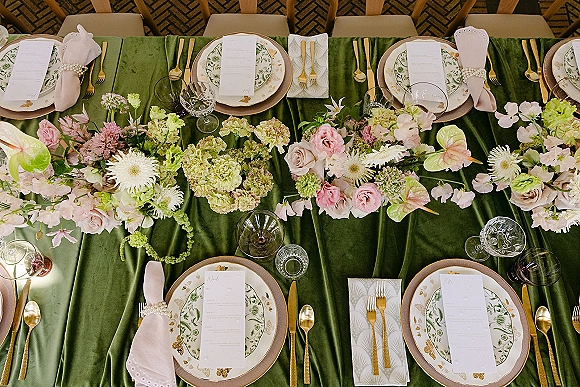 Reception tablescape with green velvet tablecloth, floral garland of roses, hydrangea and anthurium, gold flatware, and pearl napkin rings on brick floor