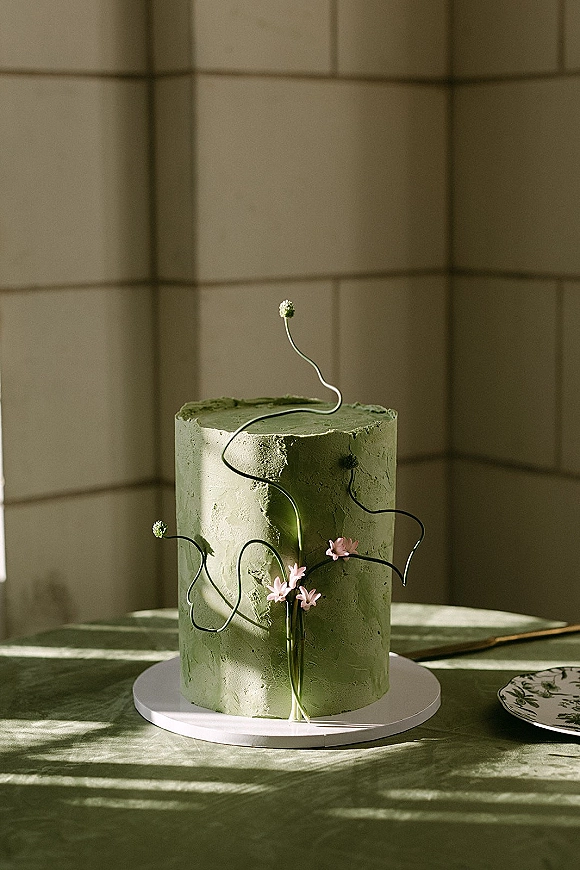 Wedding cake in sage green buttercream with sculptural floral stems and small pink flowers on a white board in soft window light